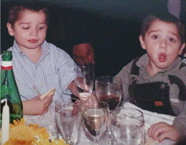 Two children at a dinner table. Multiple glasses in front of them with different heights, some containing water, others with wine. To the left, part of a composition with two big orange flowers and a candle is visibile; next to it, a green water bottle. The child on the left has a checkered powder blue shirt, and is holding what seems to be a piece of bread; his face is playful and looking at the glass of wine that he's reaching out to. The other child has a black romper on a grey shirt and is looking straight into the camera and sticking his tongue out.