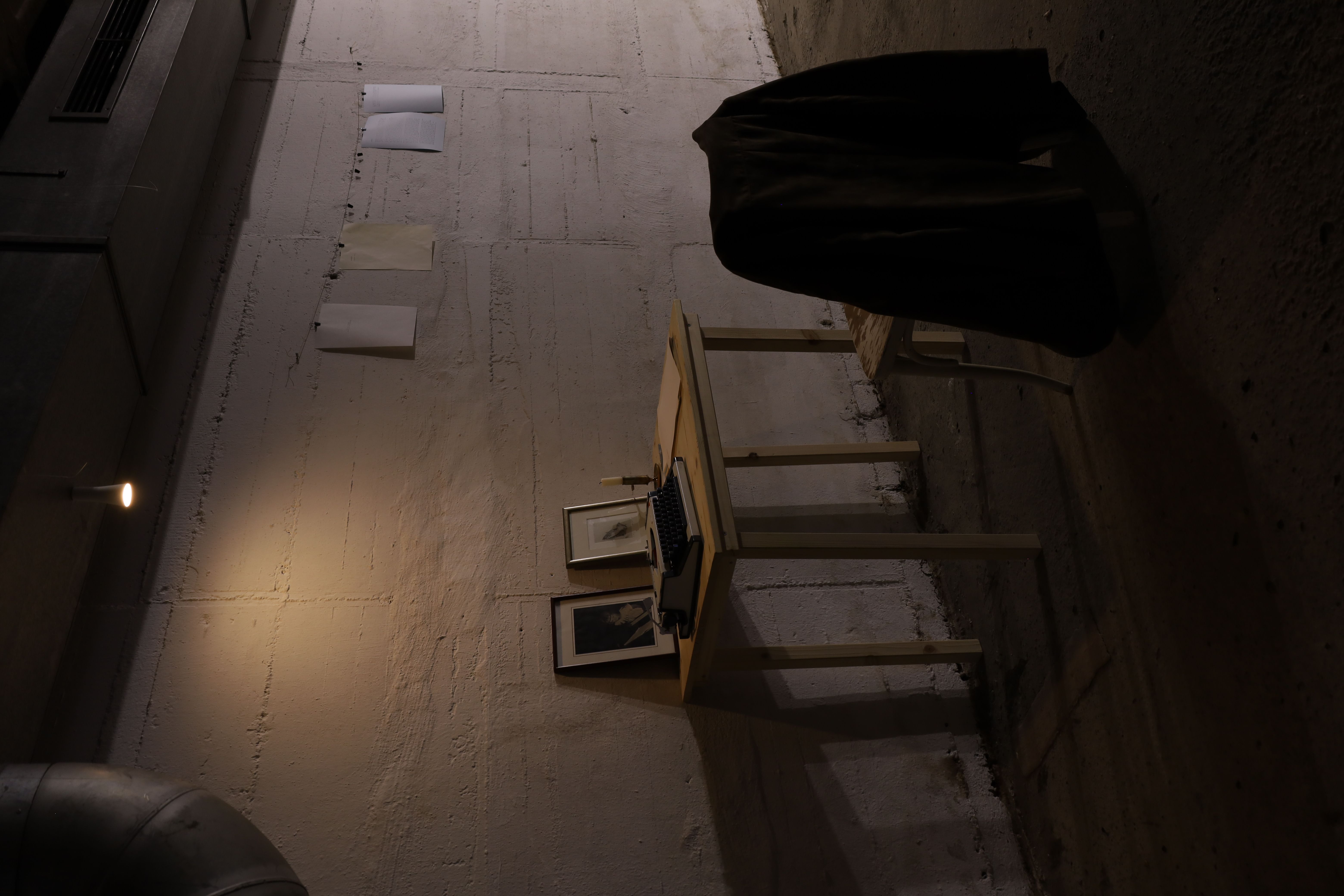 An undeground parking space with dim lighting. An overhead light illuminates a desk with a typewriter and two framed pictures, one with a child playing on piano and the other with a drawing of praying hands. In front of the latter sits a candle flame, not lighted, ina  bronze holder. Next to the typewriter are pages, which could be used by visitors in conjunction with the machine. In front of the desk is a chair with a an jacket fashioned in the traditional German style, or Tracht. Next to the desk papers are hung up on a string across the wall.